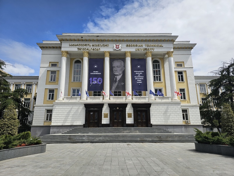 Front view of the Georgian Technical University building in Tbilisi, featuring a white facade with gold columns, large banners celebrating a 150th anniversary, and Georgian flags.