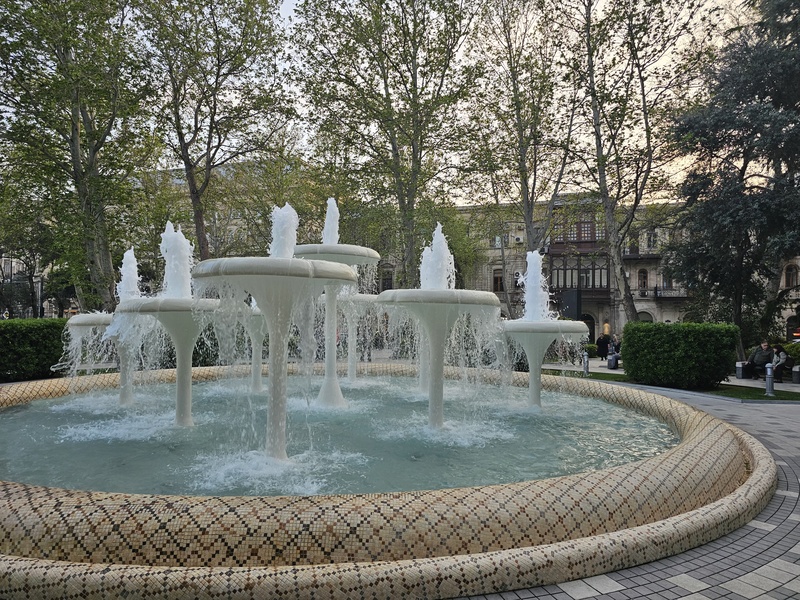 A circular fountain with multiple white mushroom-shaped jets spraying water into a tiled basin, surrounded by trees and historic buildings in Baku.
