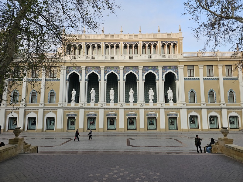 The facade of the Nizami Museum of Azerbaijani Literature in Baku, featuring white statues in arched alcoves and blue tilework, with visitors walking in front.