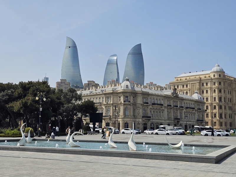 White swan sculptures in a fountain with the Flame Towers and historic buildings in the background in Baku.