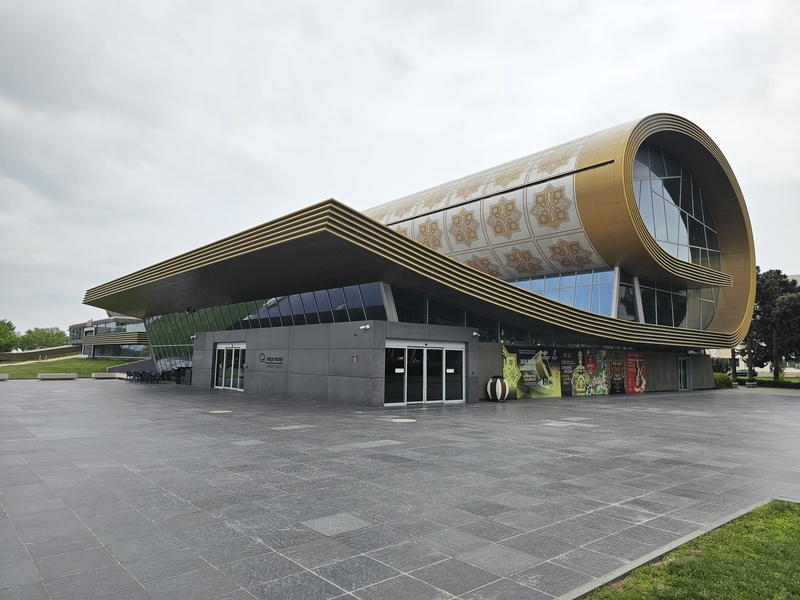 Exterior view of the Carpet Museum in Baku, featuring a modern cylindrical building with golden patterns and a large glass facade.
