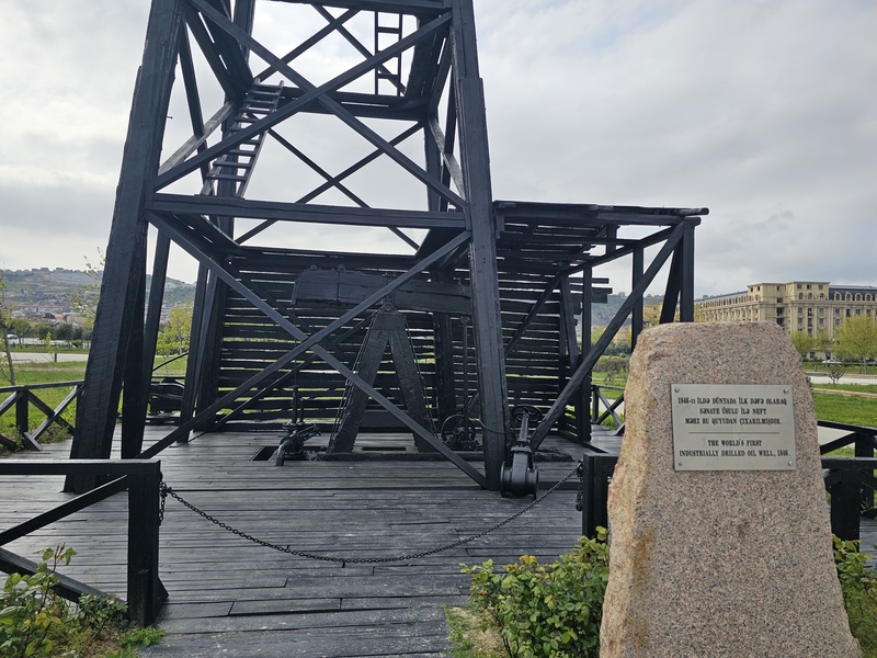 A black wooden derrick structure standing on a wooden platform, identified by a nearby stone plaque as the world's first industrially drilled oil well from 1846.