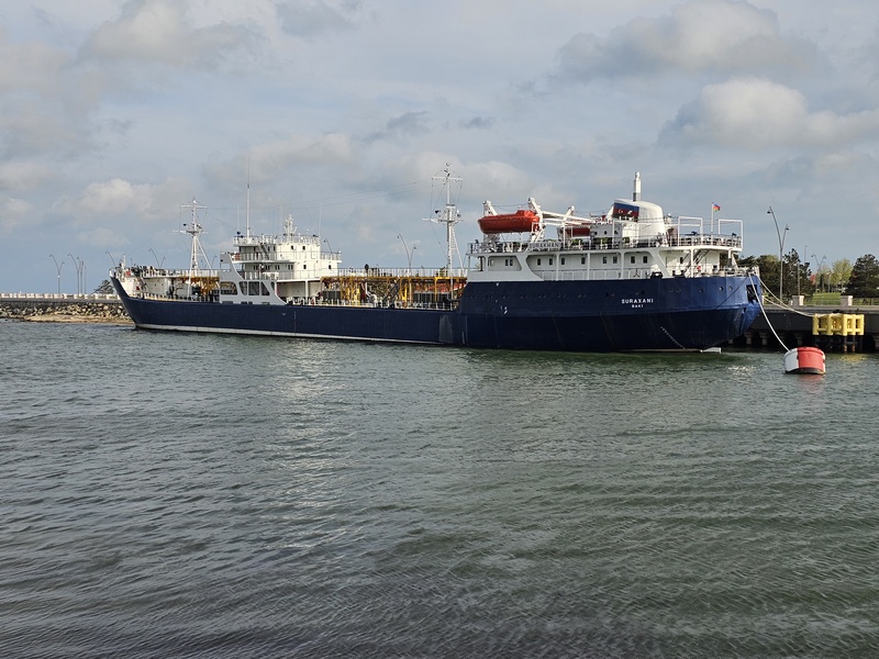 The Suraxani ship docked at the port in Baku, Azerbaijan, with a blue hull and white superstructure, under a partly cloudy sky.