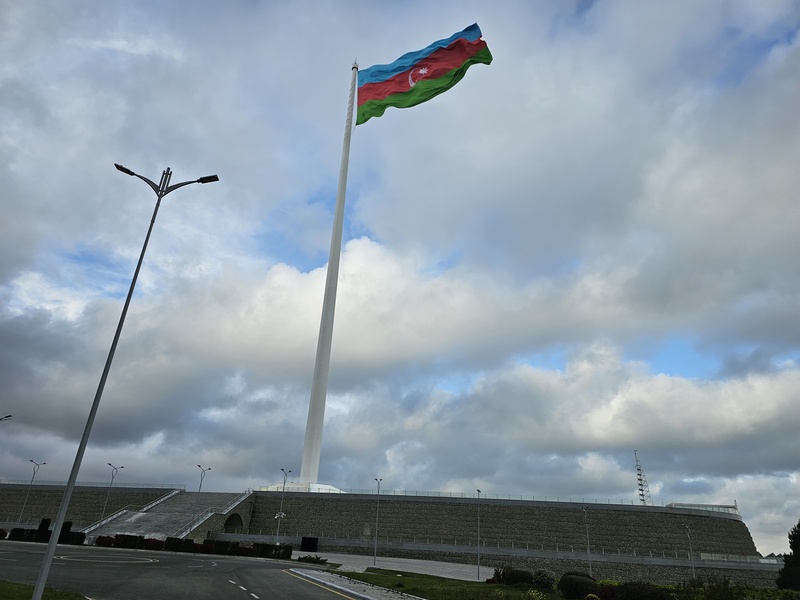A massive Azerbaijani flag waving atop a towering white flagpole against a cloudy sky, overlooking a stone retaining wall and road.