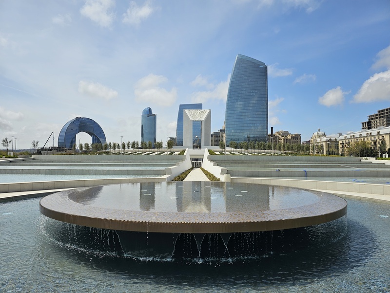 A circular fountain with cascading water in the foreground, with modern skyscrapers and a distinctive arch-shaped building in the background under a blue sky.