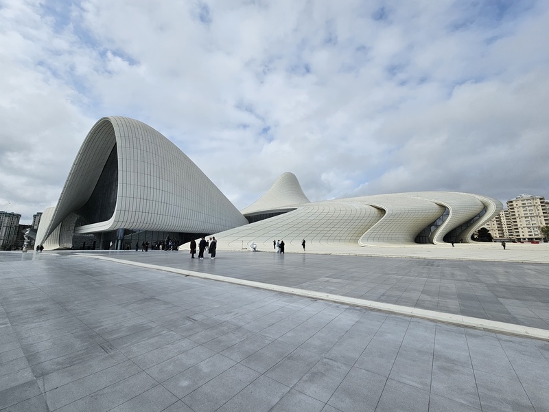 A wide shot of the white, curvilinear Heydar Aliyev Center in Baku, Azerbaijan, featuring its flowing architecture against a cloudy sky with visitors walking on the plaza.
