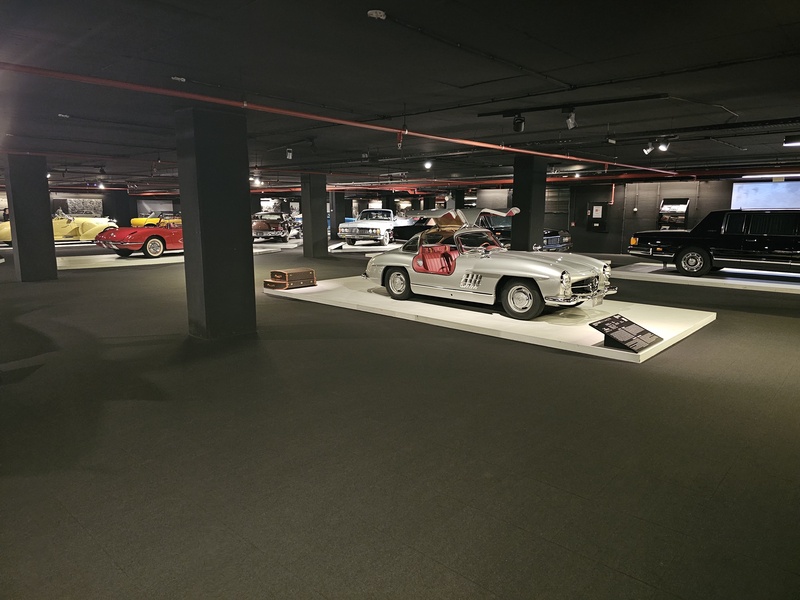 A silver Mercedes-Benz on display in a dimly lit car museum with other classic cars in the background.