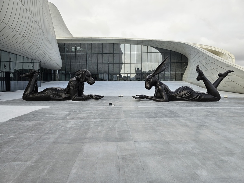 Two bronze sculptures of an anthropomorphic hare and a hound lying face-to-face on a plaza in front of the Heydar Aliyev Center in Baku.