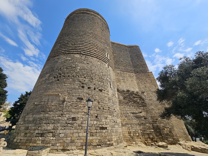 Low angle view of the ancient Maiden Tower in Baku, Azerbaijan, showing its cylindrical stone structure against a blue sky with scattered clouds.