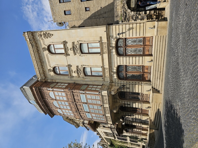 A detailed view of an ornate, historic building in Baku featuring intricate stone carvings, arched windows, and a wooden bay window under a clear blue sky.