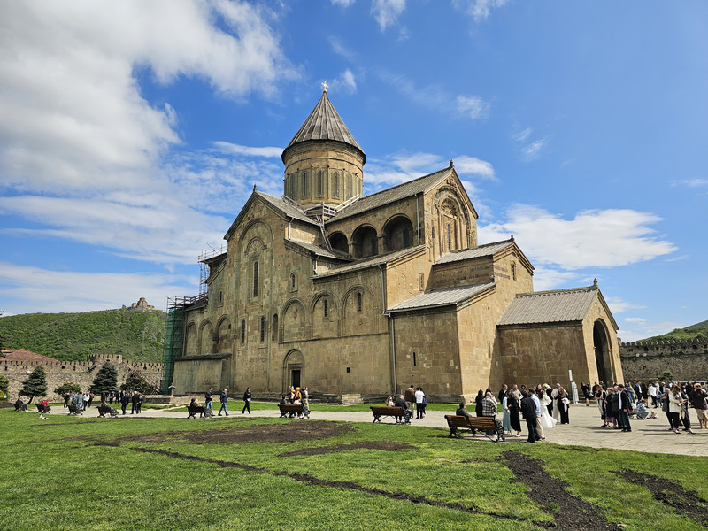 Svetitskhoveli Cathedral in Mtskheta, Georgia, with visitors on the lawn under a blue sky.