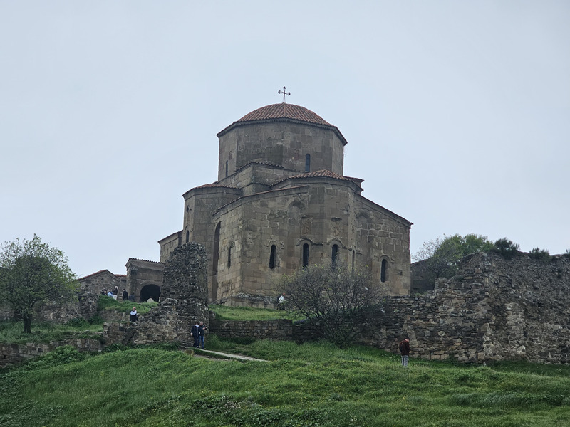 Jvari Monastery on a grassy hill under a cloudy sky.