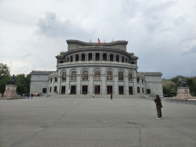 A wide shot of the grand, grey stone National Academic Theatre of Opera and Ballet in Yerevan, Armenia, featuring a large open plaza, statues on either side, and a flag flying atop the building under a cloudy sky.