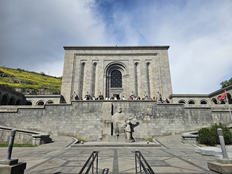 Front view of the Matenadaran in Yerevan, Armenia, featuring its massive stone facade, arched entrance, and statues of historical figures on the steps under a cloudy sky.