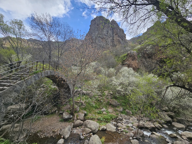 A stone arch bridge with metal railings spans a rocky stream in a lush, green valley, with a large, rugged mountain peak rising in the background under a blue sky with scattered clouds. Part of the Geghard Monastery complex.