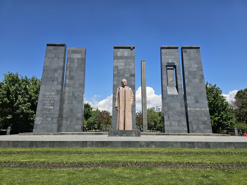 The Alexander Myasnikian Monument in Yerevan, Armenia, featuring a central statue of a man surrounded by tall grey stone pillars against a blue sky.