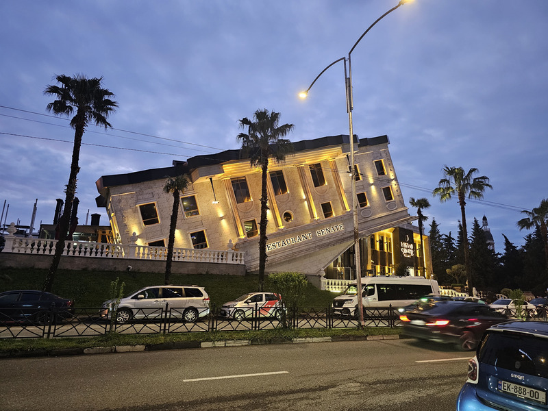 Restaurant Senate lit up at dusk with palm trees and traffic in Batumi, Georgia.