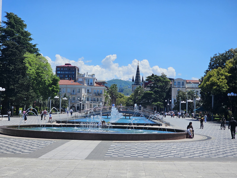 Fountains and pedestrians in a sunny Batumi plaza with trees, buildings, and mountains behind.