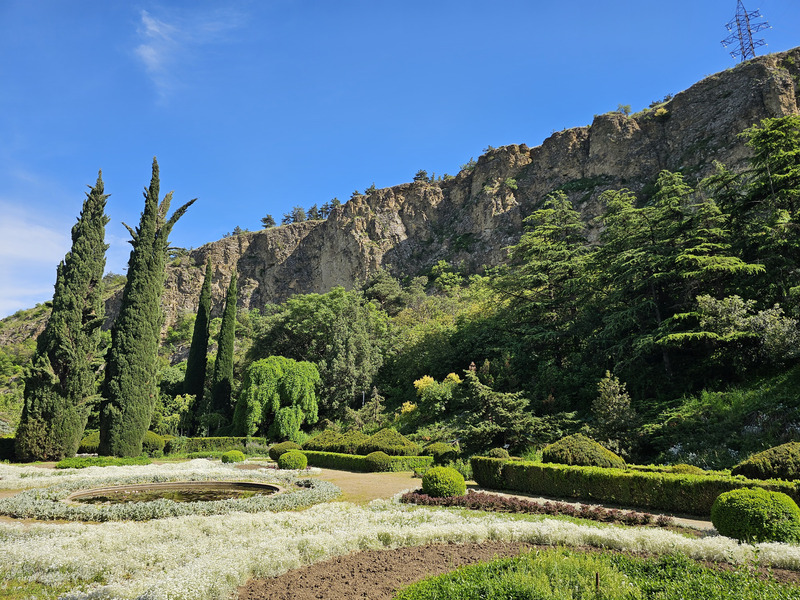 Cypress trees, flower beds, and a pond below rocky cliffs in Tbilisi Botanical Garden.