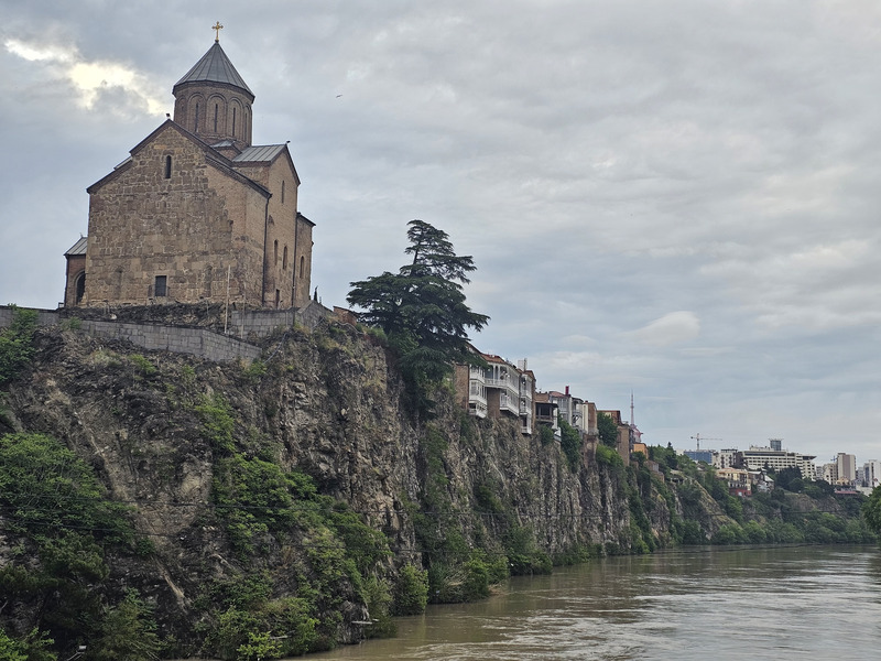 Metekhi Church on a rocky cliff above the Mtkvari River in Tbilisi under a cloudy sky.