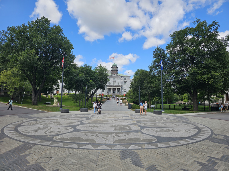 McGill University's stone Arts Building and green cupola beyond a patterned plaza, leafy trees, and a central walkway.
