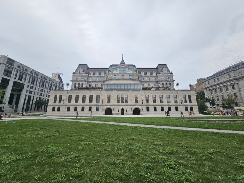 The rear of Montreal City Hall, with a green roof and rows of windows, viewed across the grass and paths of Champ-de-Mars.