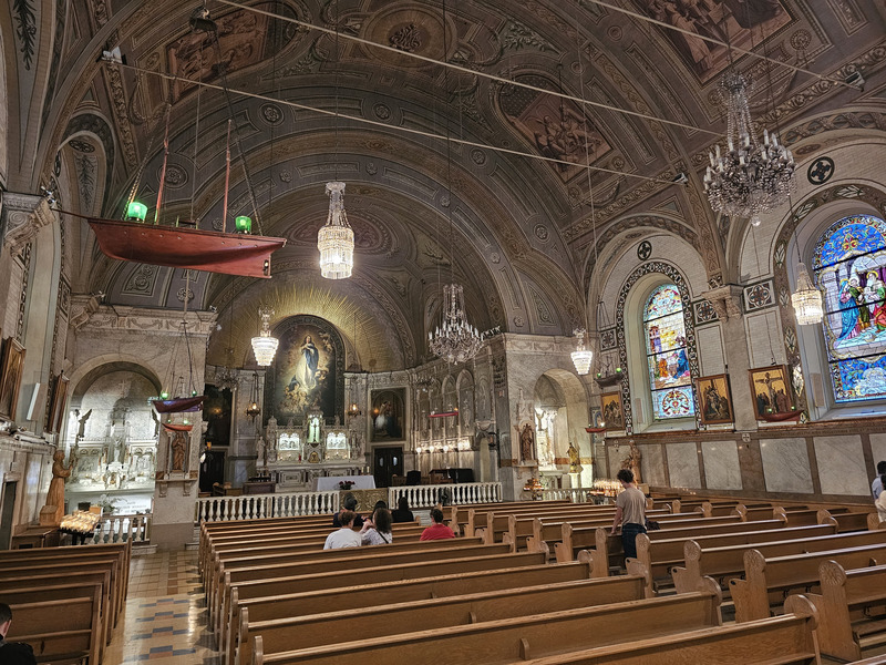 Model sailing ships and chandeliers hang above the pews in Notre-Dame-de-Bon-Secours Chapel, beside stained-glass windows and a painted vaulted ceiling.